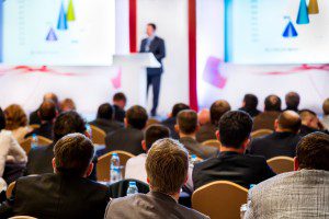 Shows a speaker at the front of an audience. Event promotion is an important part of organising a successful event.